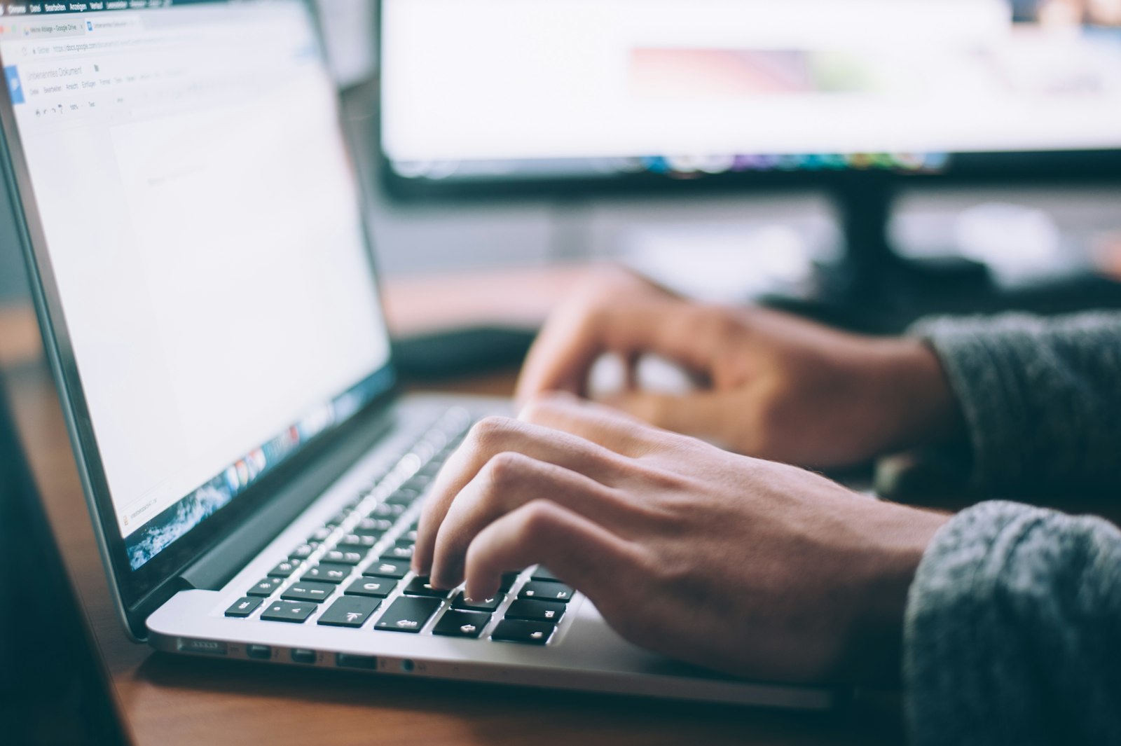 Hands typing on a laptop keyboard with a browser document open and a second monitor behind