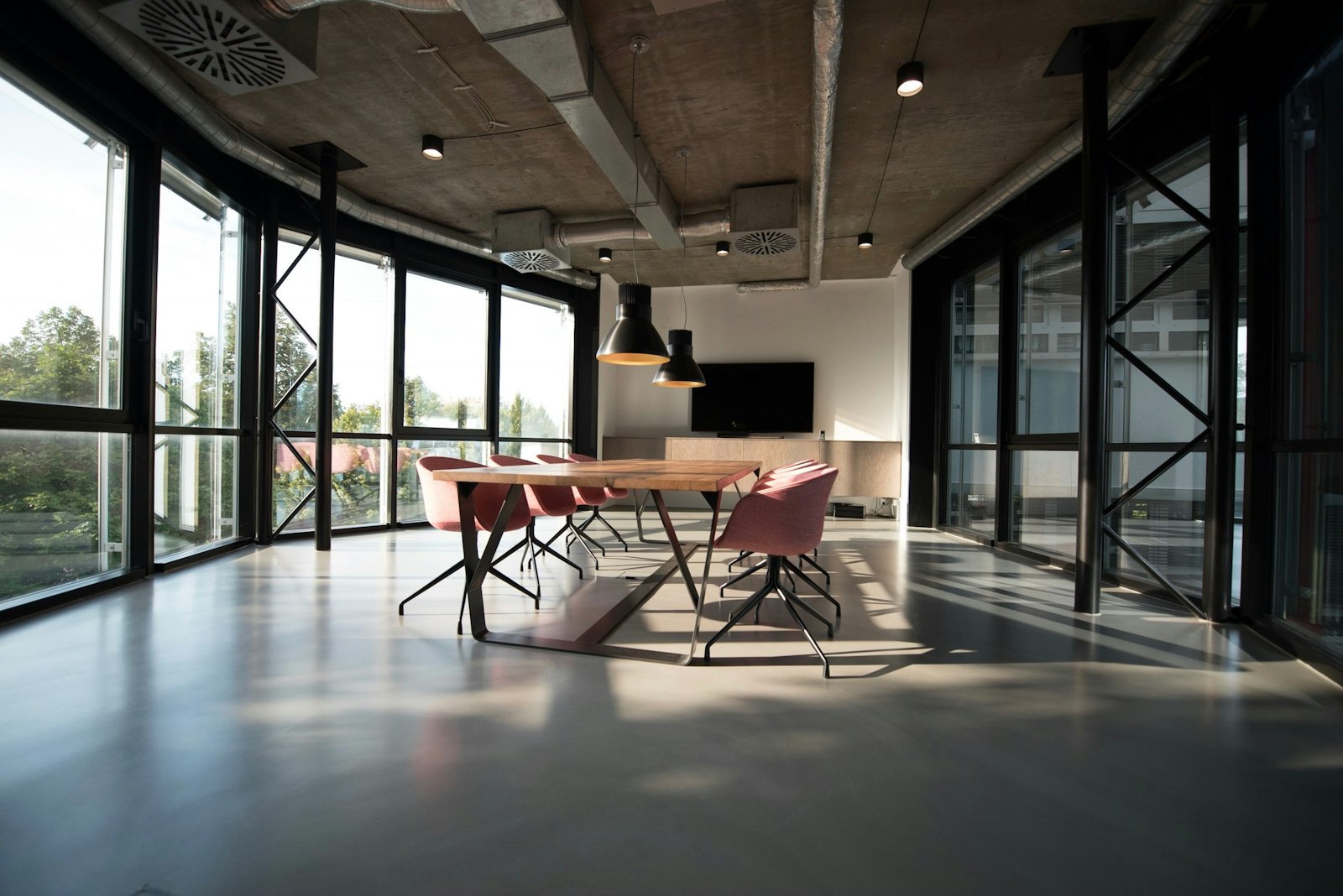 Sunlit modern meeting room with a long wooden table, pink chairs, and floor-to-ceiling glass walls