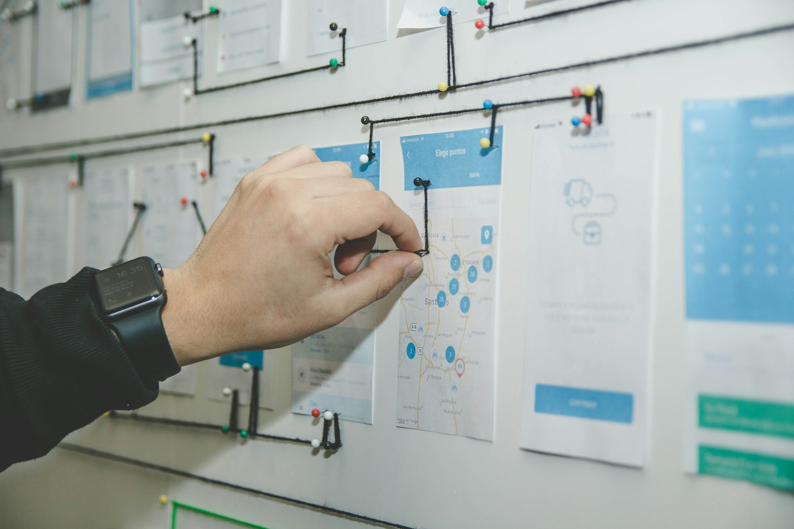 A hand pinning mobile app user-flow wireframes to a wall, connected with thread